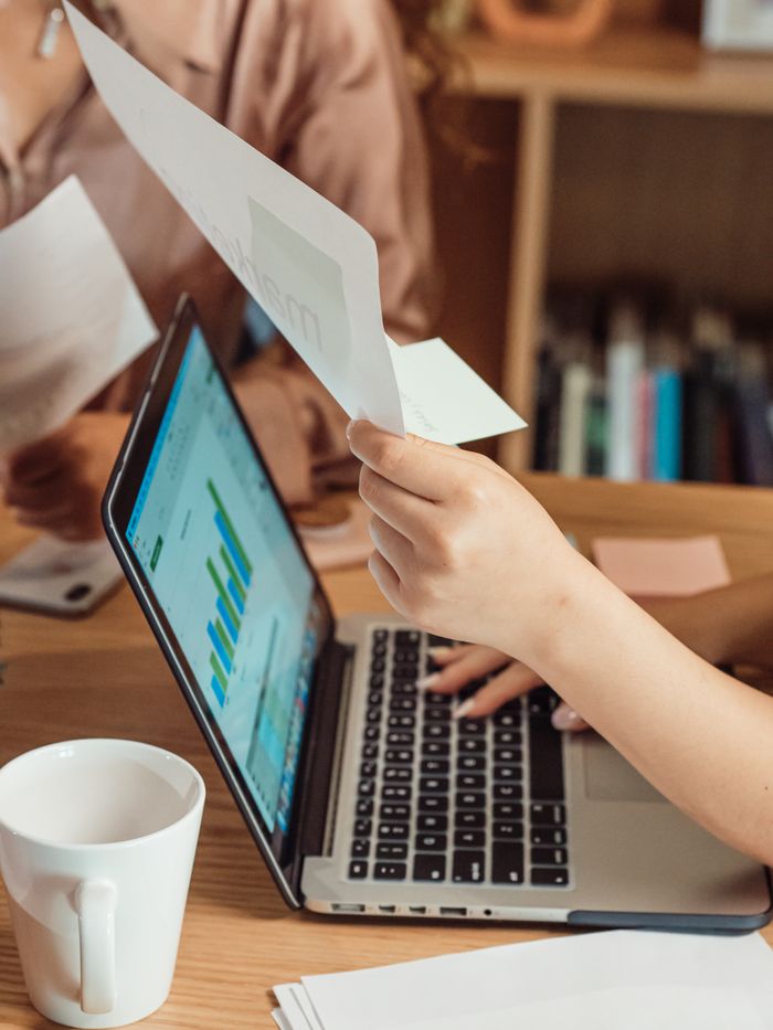 A close-up of hands holding documents over a laptop, showcasing a work setting with charts and paperworks.